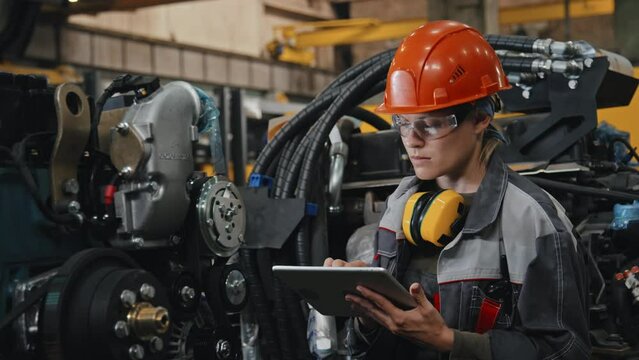 Professional female engineer in hard hat and safety glasses examining modern machine equipment at plant and taking notes on tablet computer