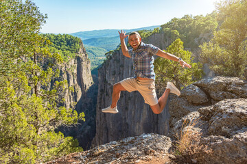 Man in a plaid blue shirt and cap jumps run smiling and shouting on the edge of deep canyon cliff,...
