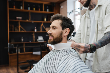 tattooed barber wrapping neck of bearded man with hairdressing collar.