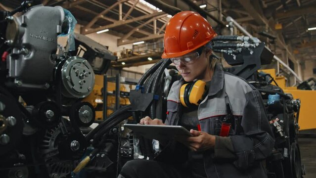 Young female engineer in safety glasses and hard hat taking notes on digital tablet while examining modern machine equipment at industrial plant
