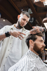Fototapeta premium Tattooed hairstylist cutting hair of bearded client in beauty salon.