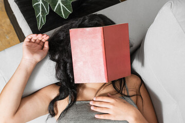 brunette girl lying on her couch with a red book placed on her face. latina woman falling asleep reading a book.