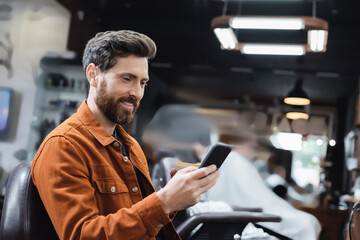 happy bearded man using mobile phone in blurred barbershop.