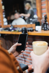 cropped view of blurred man holding smartphone with blank screen and glass of cappuccino in barbershop.