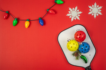 Christmas Pickleball flat lay with yellow, red and blue Pickleballs on a black and white Paddle.  Red background