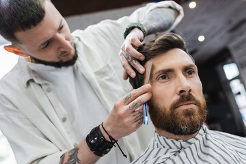 brunette bearded man near barber shaving his forehead with straight razor.