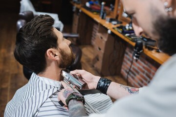 tattooed barber shaving neck of brunette man with electric razor on blurred foreground.