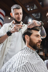 Obraz premium Low angle view of blurred barber holding scissors and comb while touching hair of client in barbershop.