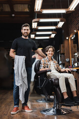 Fototapeta na wymiar Smiling hairstylist holding cape near teenager in armchair in beauty salon.