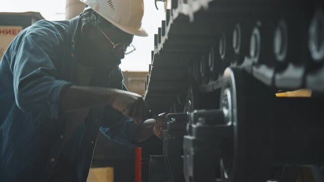 Black male service technician in denim shirt and hard hat using wrench while fixing tractor machine indoors