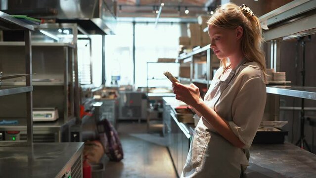 Positive blonde woman chef typing on phone in the restaurant kitchen