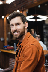 brunette man with beard smiling at camera in blurred barbershop.