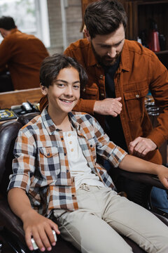 Smiling teen boy looking at camera while sitting near barber in barbershop.
