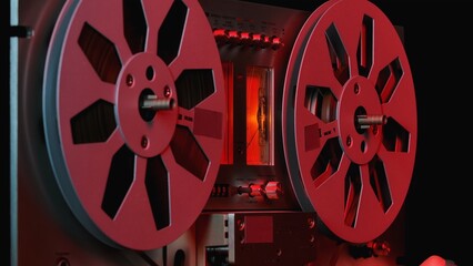 Reel to reel tape recorder on black background with red light. Vintage music player with round metallic bobbins close up. Retro magnetic tape reel. Popular disco trends 70s, 80s.