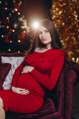 A pregnant girl is sitting on the sofa against the background of a Christmas tree