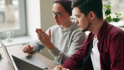 Two hipster coworkers sharing ideas about project sitting with laptops in spacious open space office. Developer discuss software program code. Designer and manager consulting on working plan.