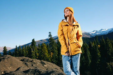 Woman with red hair hiker in yellow raincoat and cap standing on a mountain overlooking snowy mountains traveling in winter and hiking in the mountains in the wilderness at sunset