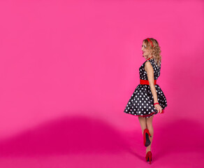 A blonde girl on a pink background in a black dress with white polka dots with red accessories: high heel shoes, beads with a bracelet and earrings, a hoop on her head. Lips with red lipstick. Pin-up.