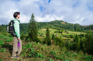 Young man hiker in a green jacket and with a backpack looks at the autumn mountains with a forest, photo of a tourist