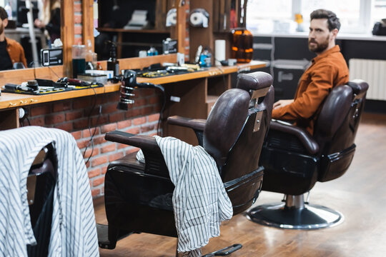 bearded man sitting in hairdressing chair and looking at camera in barbershop.