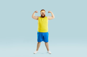 Funny happy positive cheerful excited motivated fat man in sweatband, yellow top and blue shorts smiling, flexing arms and showing his weak muscles. Sports exercise, fitness workout at the gym concept