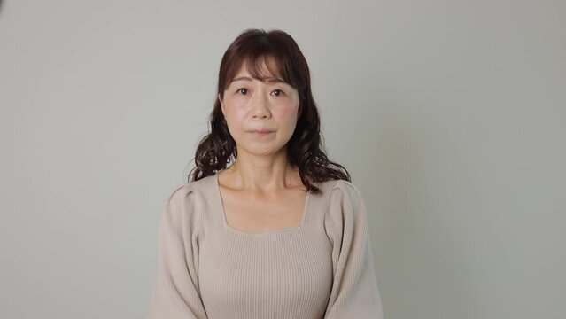 Middle aged asian woman stand still and stars the camera. White wall. Bust-up