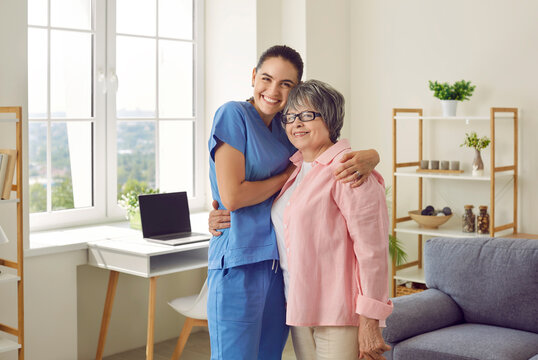 Happy Beautiful Senior Woman Together With Her Friendly Young Nurse Or Caregiver Standing In Room In Retirement Home, Hugging Each Other, Looking At Camera And Smiling