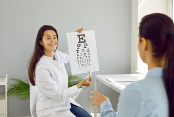 Optical eye test in oculist office. Ophthalmologist checks woman's eyesight by pointing to small letters on chart for vision test. Friendly smiling young female doctor holding Snellen chart.