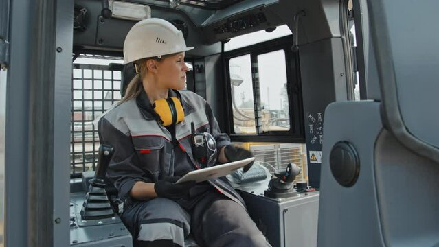 Young Caucasian female engineer in hard hat and coverall setting up parameters in tractor cab using digital tablet