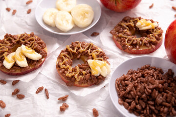 Obraz na płótnie Canvas Creative party snack for holidays. Apple rounds with peanut butter, caramel and chocolate flavor puffed rice topping with banana slices. Funny appetizer for kids and adults.