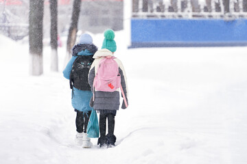 Two elementary schoolgirls go to school on a snow-covered path among snowdrifts. Snow is falling....