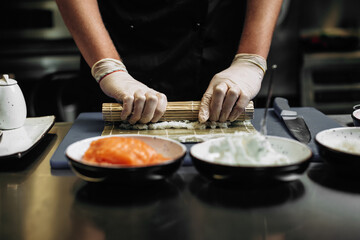 A male chef makes sushi and rolls from rice, red fish and avocado. White gloves. Dark background.