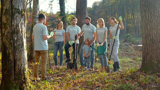 Male leader of ecological activists standing with document or plan in hands and giving commands to people with kid for planting trees in forest. Outdoors. Enviromental concept. Working as volunteers.