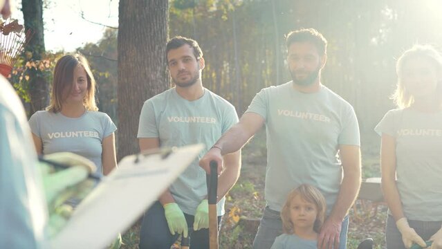 Team of Caucasian young happy volunteers with kid tanding in forest and communicating with smile. Sharing tasks for volunteering in wood. Environmental activists. Teamwork.