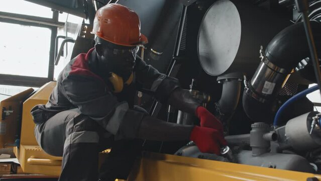 Black male mechanic in hard hat and coverall repairing pipelayer machine with wrench bolt at service centre