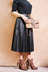 a woman wearing a skirt and holding a handbag in her right hand, standing against a beige wall background