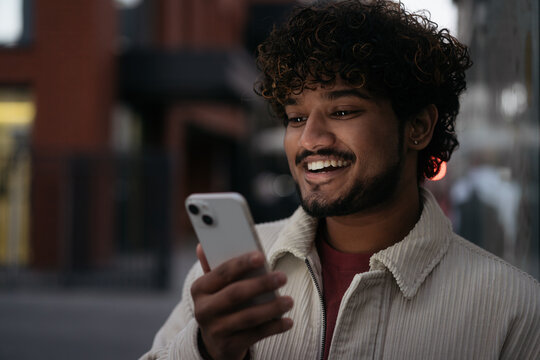 Night Portrait Of Stylish Smiling Indian Man Using Mobile Phone Reading Text Message On The Street. Asian Hipster Guy Shopping Online, Looking At Digital Screen, Selective Focus. Mobile Banking  