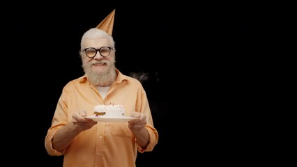 Smiling senior man blowing out candles on birthday cake on black background