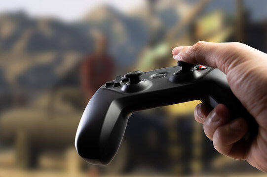 Close-up. A modern GAM22ICB gamepad in a gamer's hand against the backdrop of an adventure video game on a large plasma TV screen. Online games with friends, communication.