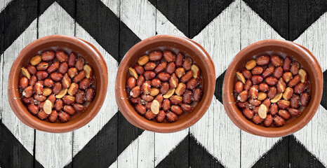 Three clay dishes of fried peanuts with skin, on a black and white painted wooden background.