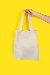 Mano de mujer sosteniendo una tote bags de algodón crudo sobre un fondo amarillo brillante liso y aislado. Vista de frente y de cerca. Copy space