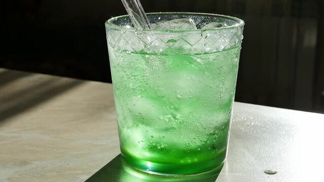 A glass filled with a green fizzy drink with ice cubes on the surface and a glass reusable drinking straw. the drink sizzles and bubbles in the sun standing on the table close-up. tarragon
