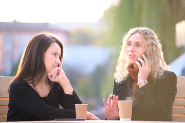 Happy young woman chatting on mobile phone ignoring her bored friend. Female friends sitting at...