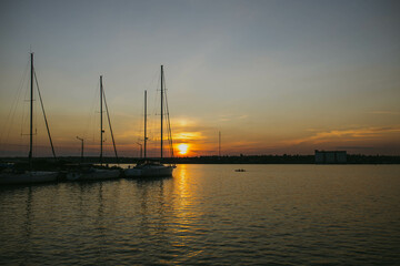 South Ukraine, Mykolaiv - August 21, 2021: Sunset in the river coast side, sun reflects into the water surface. Yachts and boats near the beach, people resting, Calm atmosphere in peaceful  city