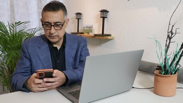 Hombre ejecutivo latino con lentes usando celular frente a computadora
