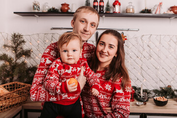Happy cheerful family celebrating New Year or Christmas at home. Young small kid with father and mother dressed red warm sweaters, spend time together smiling, standing in modern kitchen

