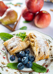 A traditional piece of apple strudel with powdered sugar, mint, blueberry and cinnamon in the foreground on a table.