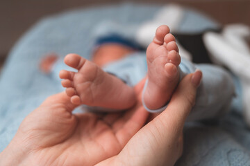 A close-up, high-angle shot captures a parent's cupped hands holding the small feet of their newborn baby. The baby is out of focus in the background, wrapped in a blue blanket.