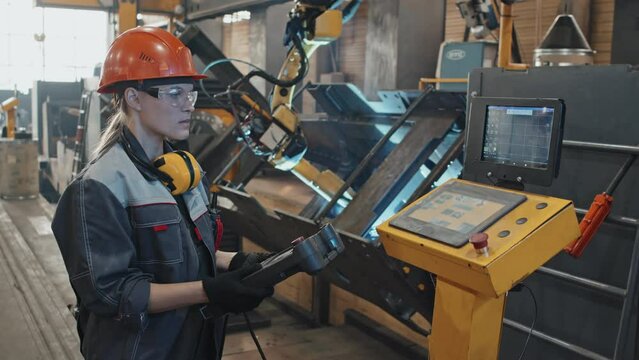 Female plant worker in hard hat and safety glasses using teach pendant robotic manipulator while operating CNC machine at work