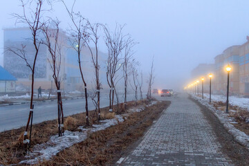 Fototapeta na wymiar Evening urban landscape. View of the sidewalk and street lights. Mid-May in the Arctic. Snow is melting on the streets of the city. Cold fog. City of Anadyr, Chukotka, Siberia, Russia.